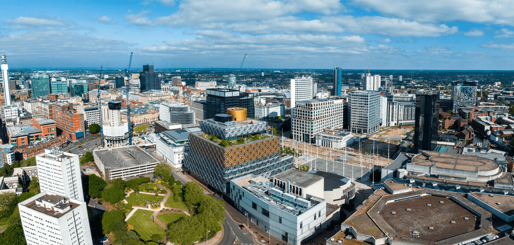 Aerial view of Birmingham city skyline with modern buildings, construction cranes, and green spaces under a blue cloudy sky.