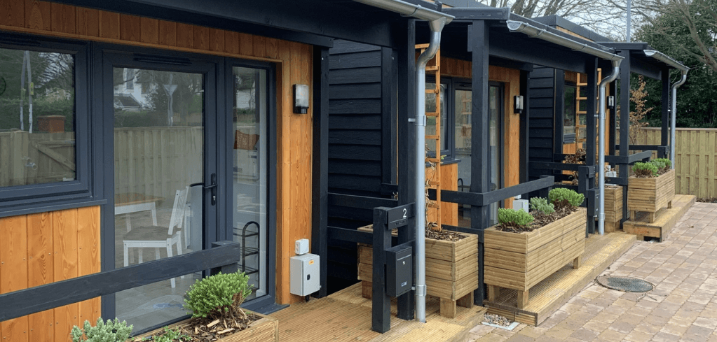 Modern wooden cabin-style units with dark trim, large glass doors, wooden decking, and planter boxes with shrubs outside.