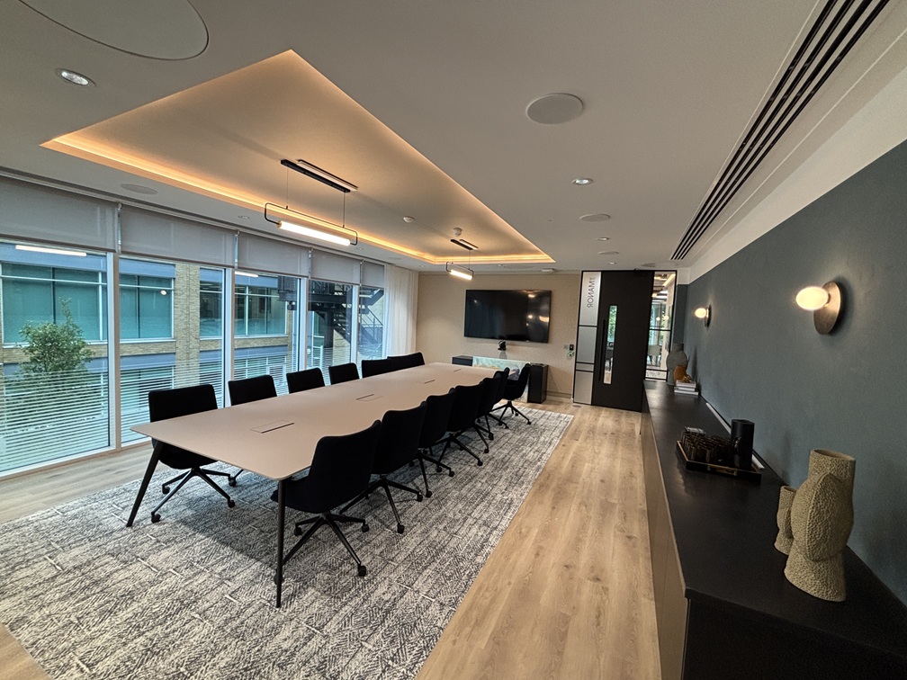 Modern conference room with long table, black chairs, wall-mounted TV, warm ceiling lighting, and large windows.