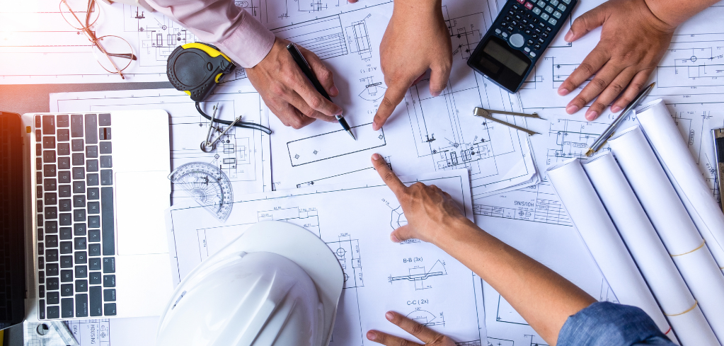 Hands reviewing architectural blueprints with measuring tools, calculator, laptop and hard hat on a workspace.
