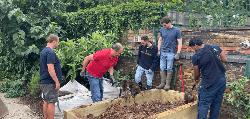 Group of volunteers working together on a garden project, filling a wooden raised bed with soil in a brick-walled garden area.