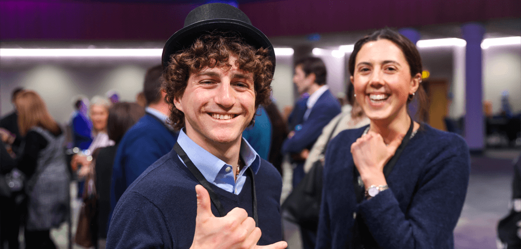 Two smiling people at a formal event, one wearing a black hat and navy sweater giving thumbs up in a crowded venue.