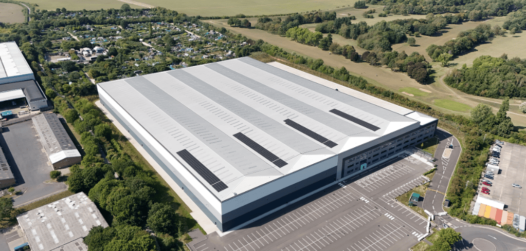 Aerial view of a large industrial warehouse with silver roof panels, surrounded by parking lots and rural countryside.