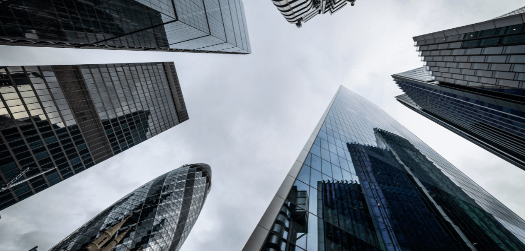Looking up at modern glass skyscrapers in a financial district, with reflective facades against a cloudy sky.