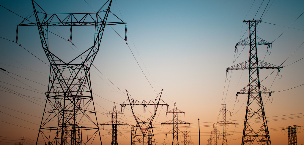 Silhouettes of electrical transmission towers and power lines against a gradient sunset sky in blue and orange.