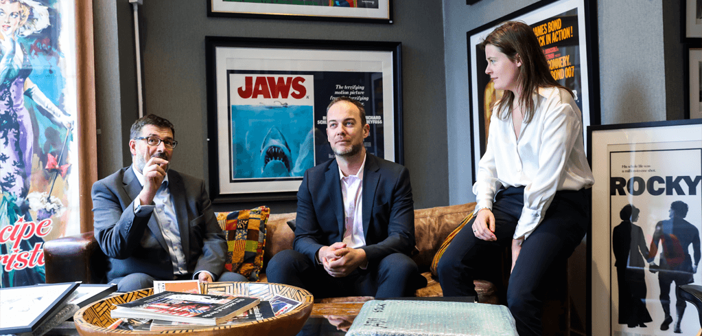 Three people in a room with classic movie posters including Jaws and Rocky displayed on gray walls behind them.