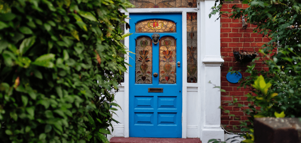 A bright blue front door with ornate stained glass panels on a red brick home, framed by lush green foliage.