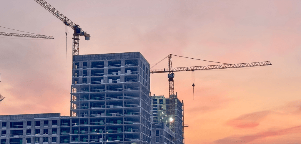 Buildings under construction with two tower cranes against a pink and orange sunset sky.