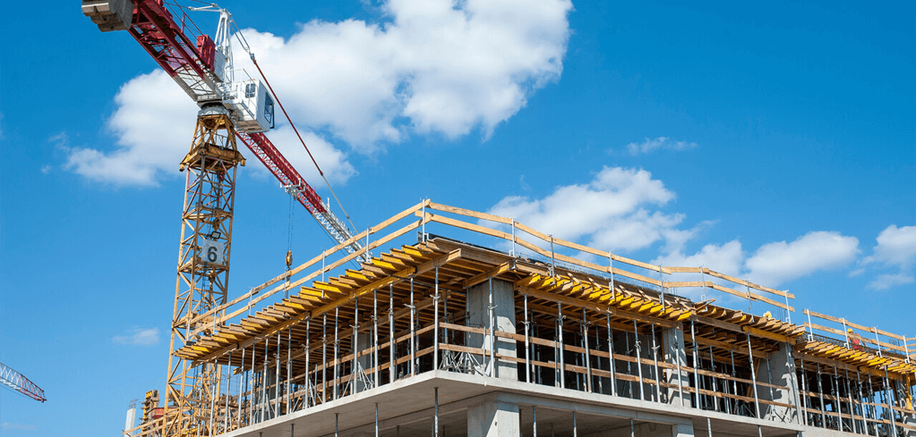 Construction site with tall crane extending over a concrete building structure against a bright blue sky with clouds.