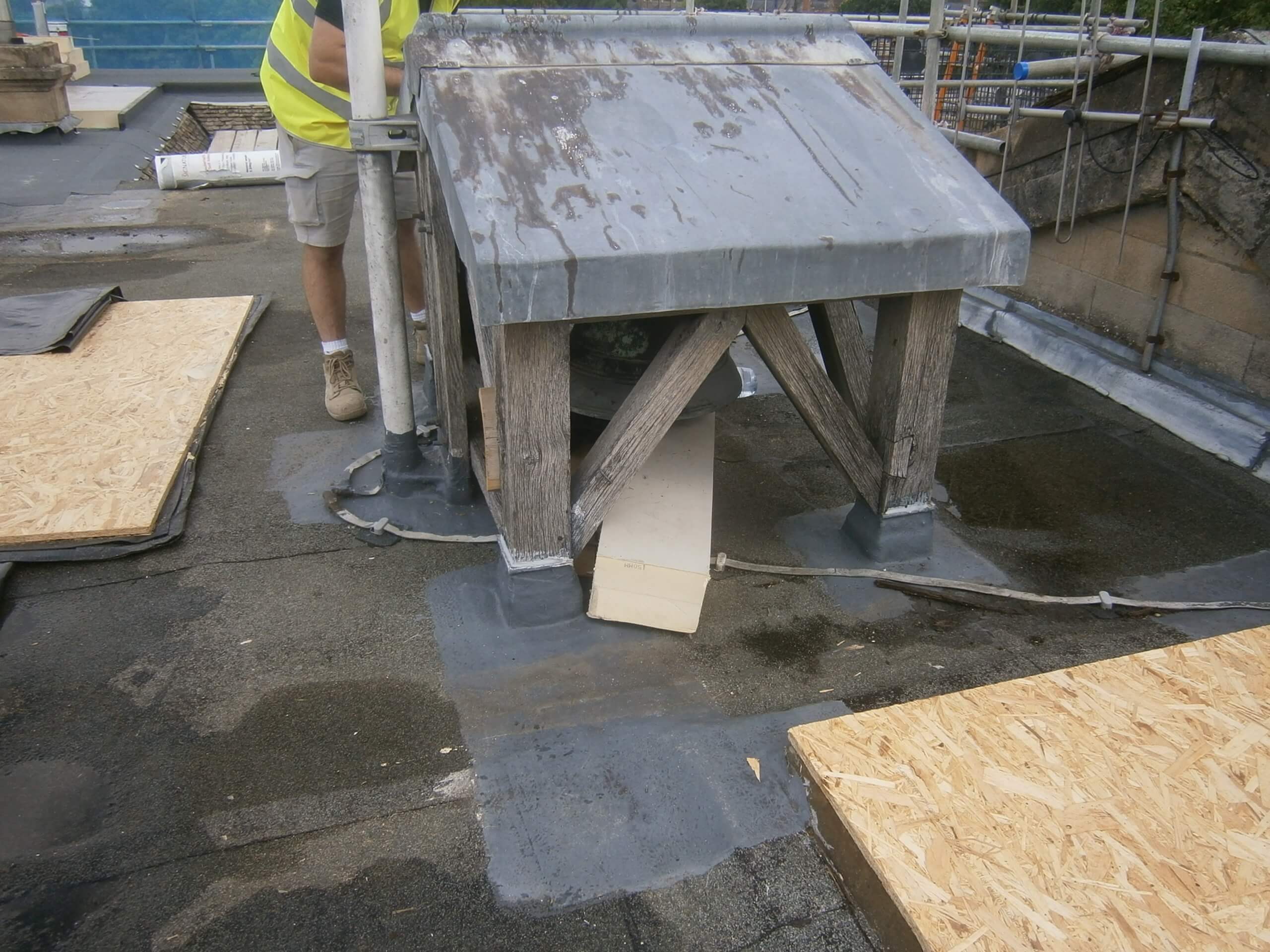 A construction worker in a yellow vest works on a flat roof near a weathered wooden structure with a metal cap.