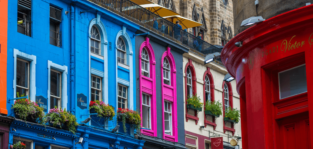Colorful storefronts on Victoria Street, Edinburgh, featuring vibrant blue, pink, and red facades with flower boxes.
