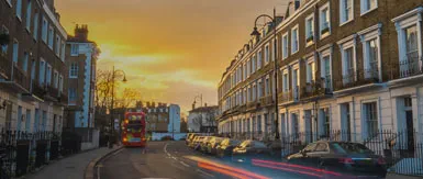 A wide street scene at sunset featuring elegant Georgian-style terraced townhouses, parked cars along the curb, and a classic red double-decker bus passing through. Warm golden light fills the sky while subtle light trails from traffic add a sense of movement, creating a calm yet lively urban atmosphere typical of a London neighbourhood.