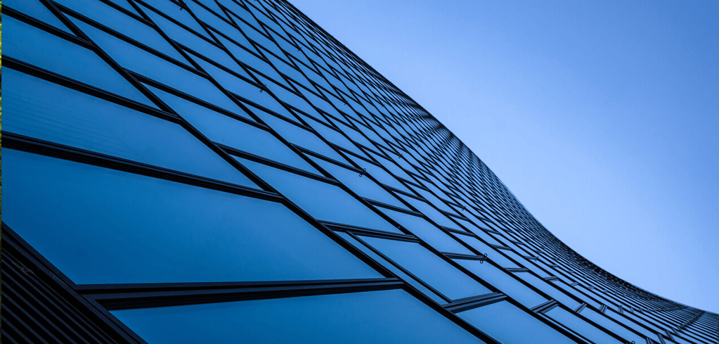 Modern glass building facade with blue reflective windows curving against clear sky, viewed from below.