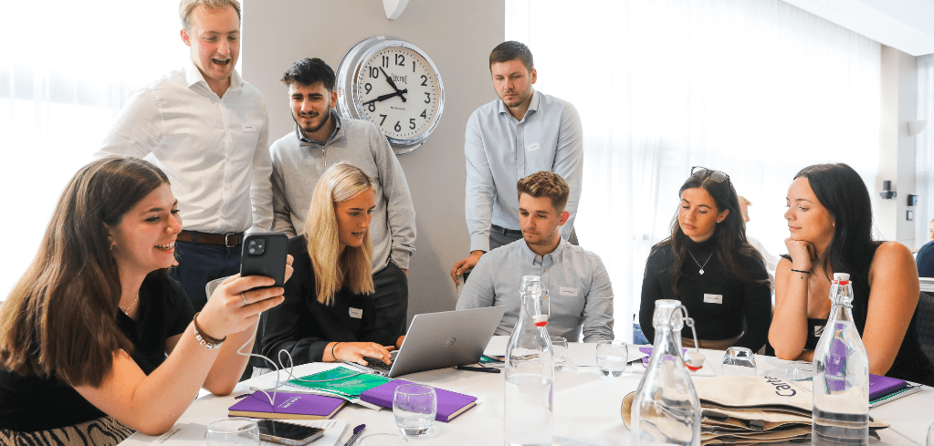 Business team gathered around table with laptop, looking at phone, in bright office with wall clock showing 9:00.