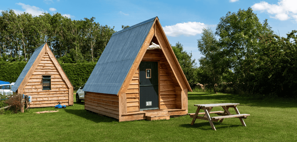 Two wooden A-frame cabins with blue roofs on green grass, surrounded by trees, with a picnic table nearby.