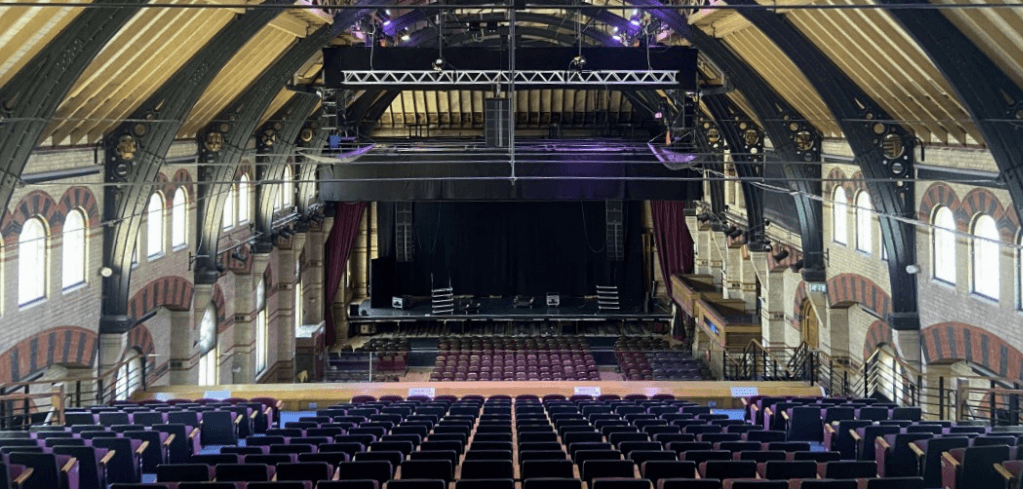 Interior of a historic concert hall with arched brick walls, wooden ceiling, stage lighting rigs, red curtains, and rows of empty seats.
