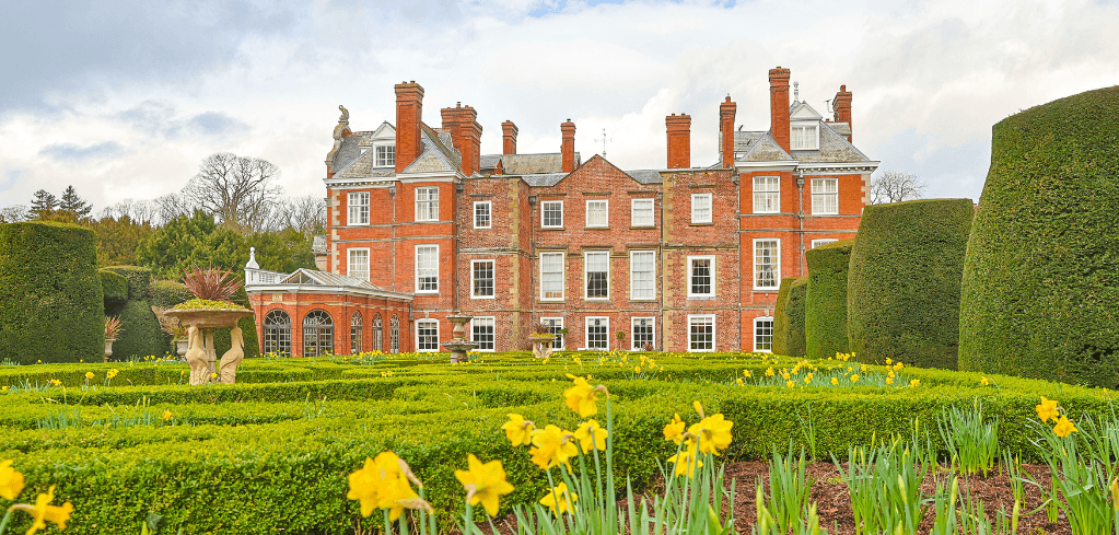 A grand red brick English manor with manicured hedges, topiary, yellow daffodils, and a stone fountain in a formal garden.