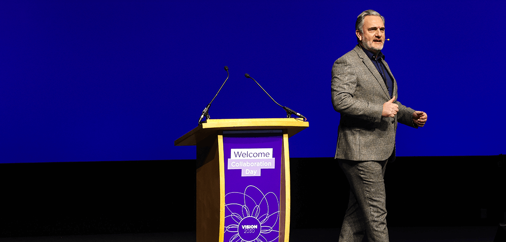 Speaker in gray suit presenting at a podium with "Welcome Collaboration Day" sign against a blue background.