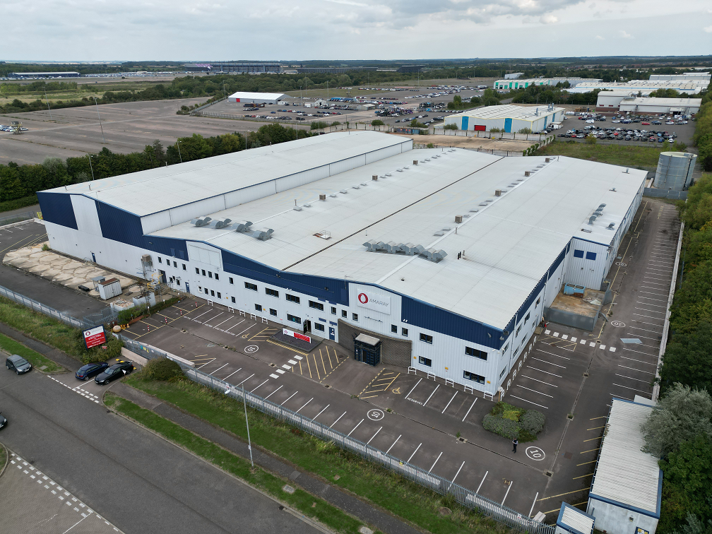 Aerial view of large industrial warehouse with white roof and blue trim, surrounded by parking lots in an industrial park.