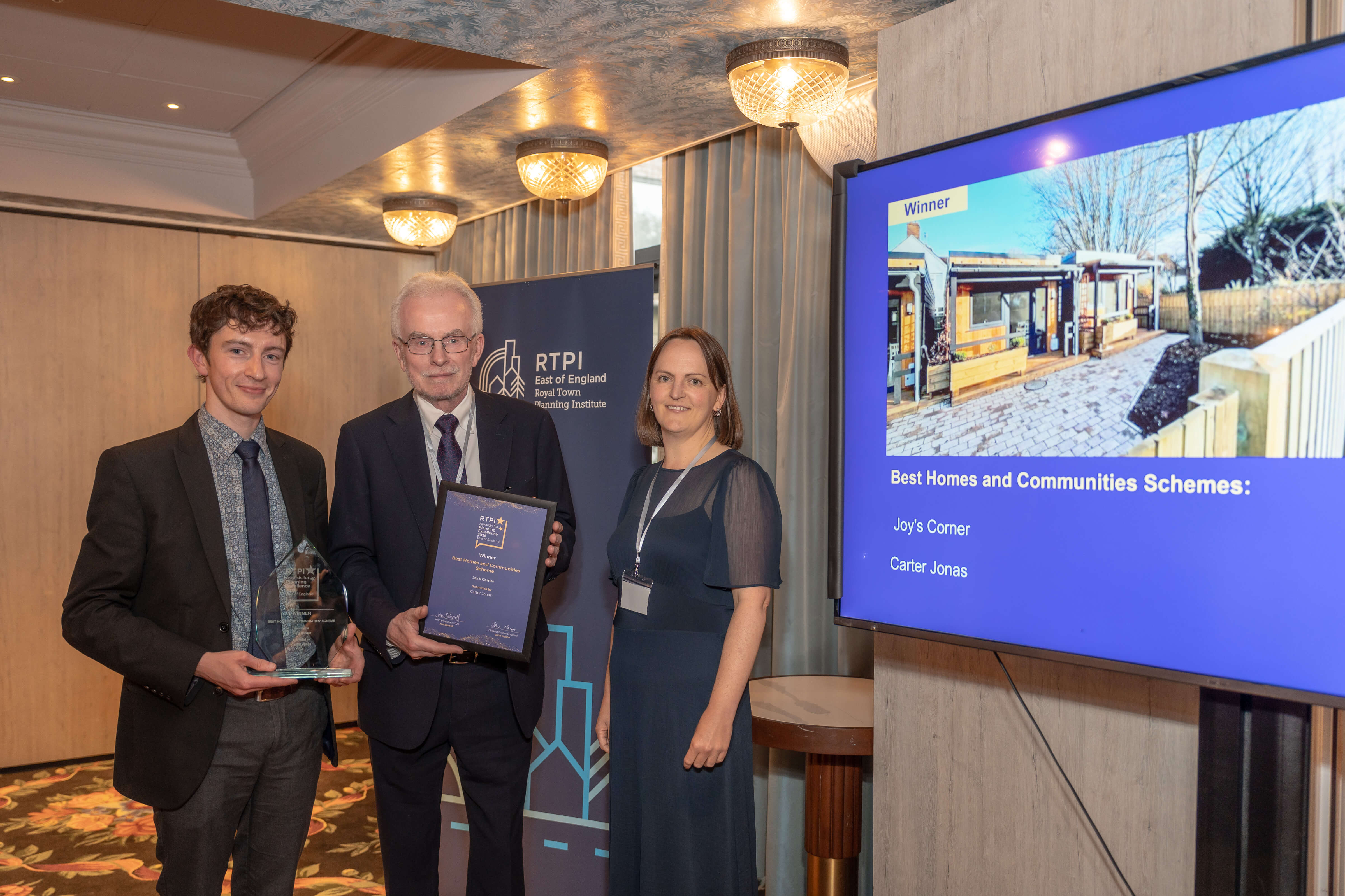 Three people pose with RTPI awards at an event, next to a screen displaying the Best Homes and Communities Schemes winner, Joy's Corner.