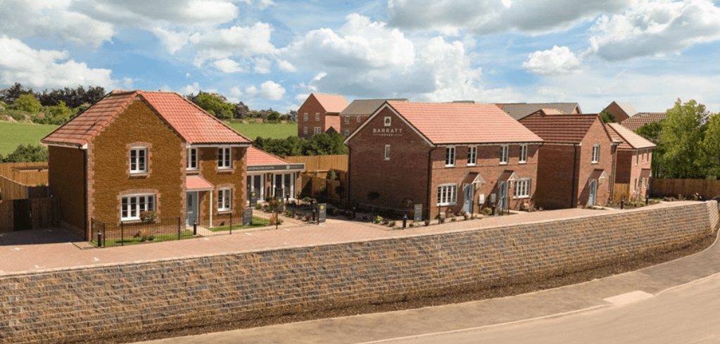 Barratt Homes new-build brick houses with red tile roofs, set behind a stone wall on a sunny day with green hills.