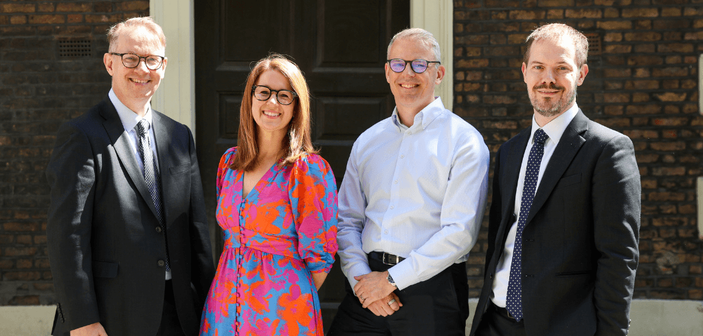Four smiling professionals, three men in suits and a woman in a colorful floral dress, standing outdoors in front of a brick building.