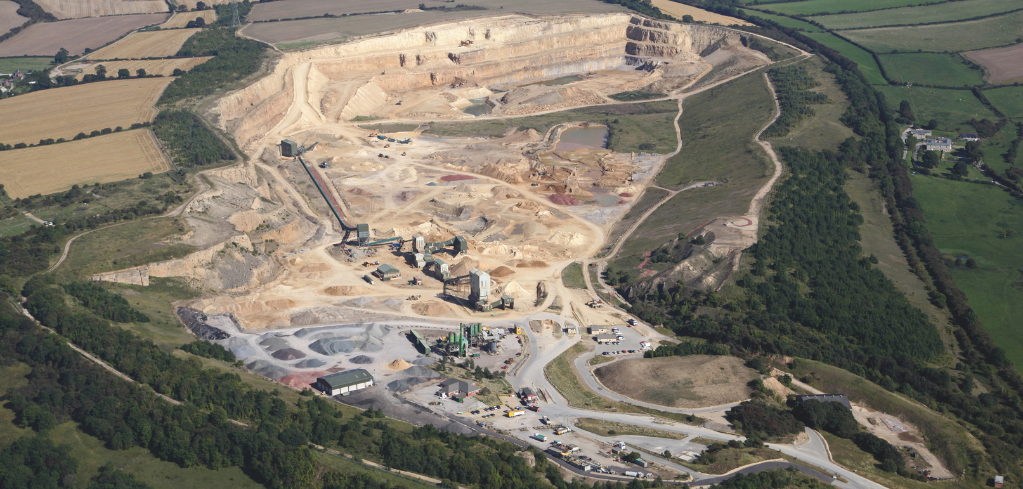 Aerial view of a large quarry or mining operation with terraced excavation levels, surrounded by farmland and green fields.