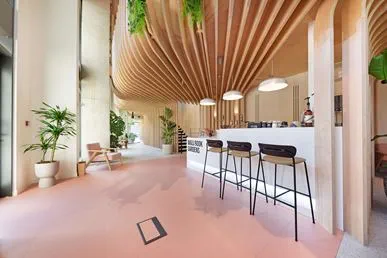 This image showcases a modern, design-led interior featuring a sleek reception or café-style counter with contemporary bar stools and soft ambient lighting. The curved wooden ceiling slats and warm pastel flooring create a welcoming, architecturally sophisticated atmosphere. Lush indoor plants and minimalist furniture enhance the sense of calm, making the space ideal for community living, shared amenities, or premium residential developments.