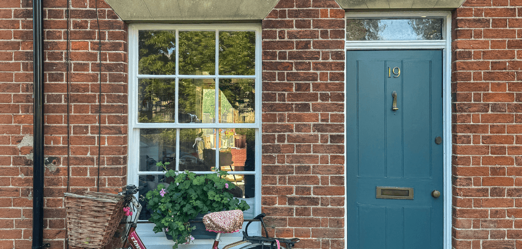 Brick house with teal blue door numbered 19, white-framed window, and partial view of a bicycle with basket in foreground.