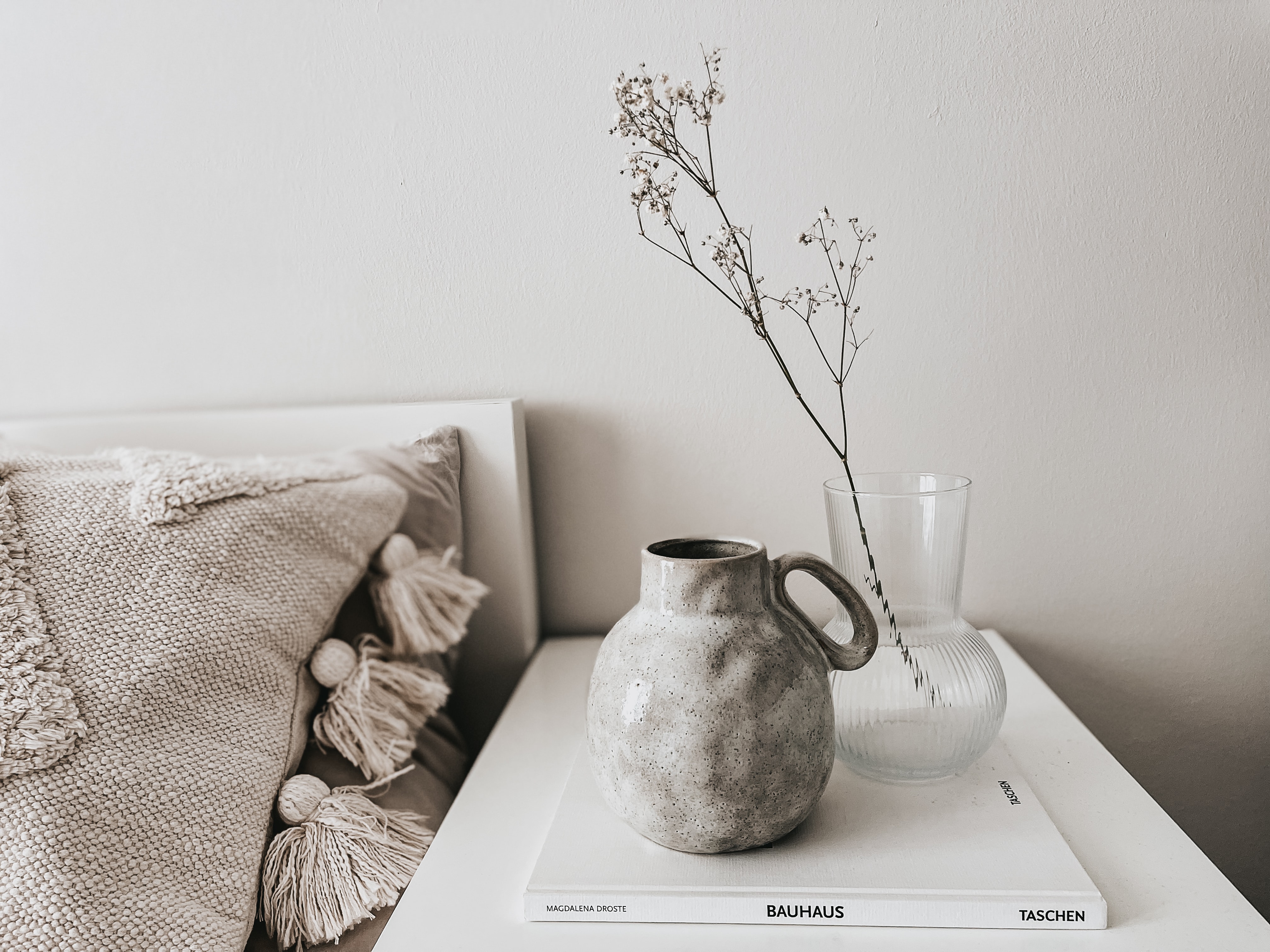 Minimalist home decor with ceramic vase, glass vase with dried stems, and Bauhaus book on white table beside textured pillows.