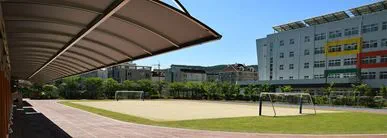An outdoor school playing field with two football goalposts, surrounded by modern buildings and greenery under a bright blue sky, with a covered walkway in the foreground.