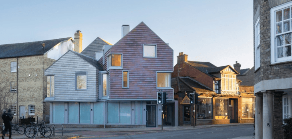 A modern timber-clad building with a pointed roof stands among traditional brick and stone buildings on a quiet street.