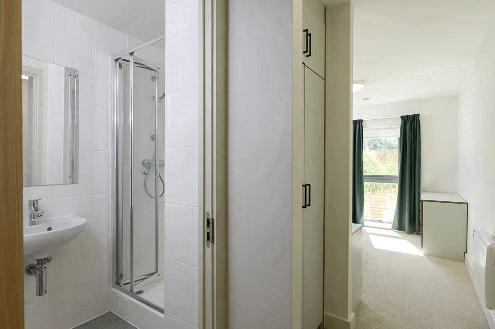 Modern apartment interior showing a white bathroom with shower stall and sink, connected to a bright bedroom with green curtains.