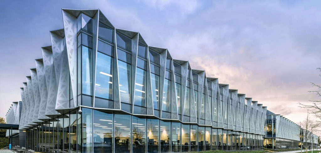 Modern glass and steel building with dramatic angular fin-shaped facade panels, illuminated interiors, under a dusk sky.
