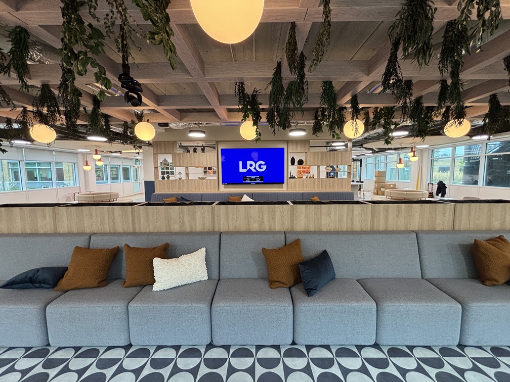 Modern office lounge with gray sectional sofa, hanging plants, round yellow lights, and large blue LRG display screen.