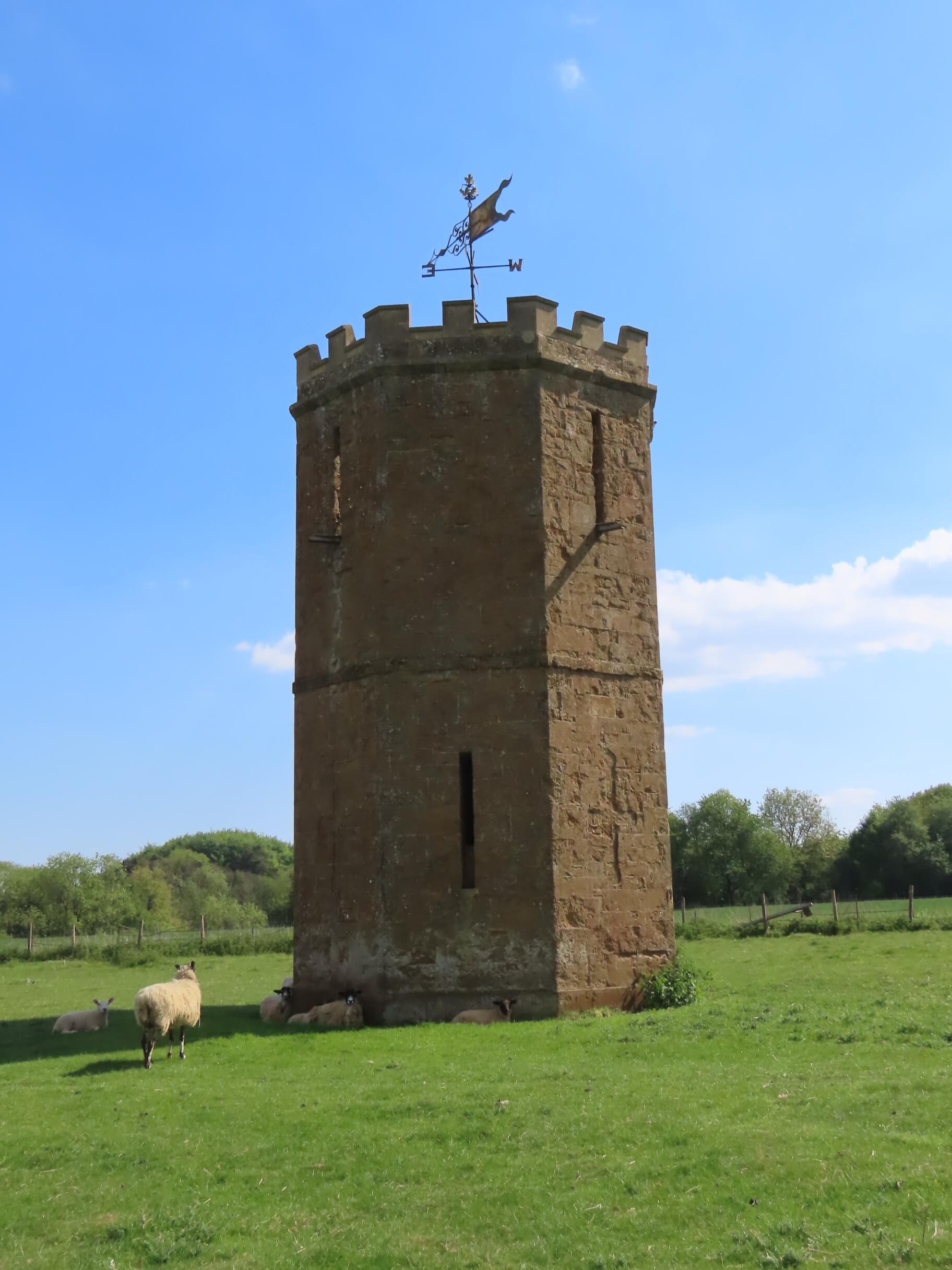A historic stone tower with battlements and a weather vane stands in a green field with sheep resting nearby under a blue sky.