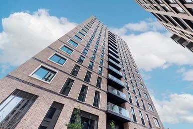This image showcases a contemporary high-rise residential tower viewed from a low angle, emphasizing its height, clean brick façade, and modern architectural detailing. The large windows and private balconies highlight premium urban living, while the bright blue sky adds a sense of openness and quality. 
