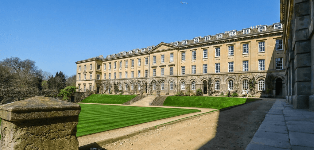 Historic stone university building with manicured green lawn, stone steps, and clear blue sky above.