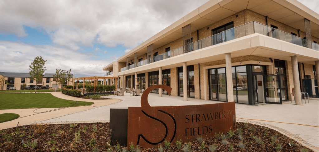 Exterior of Strawberry Fields building, a modern two-story tan brick structure with balconies, landscaped grounds, and a rustic metal sign.