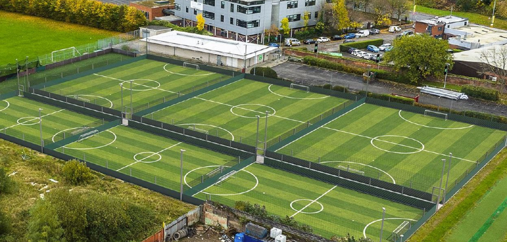 Aerial view of multiple green soccer fields with white markings, surrounded by fencing and adjacent to residential buildings.