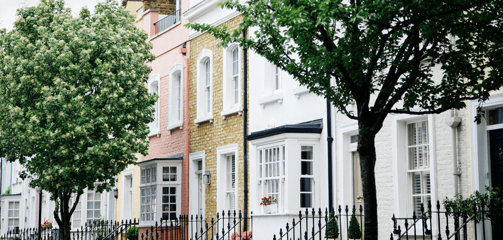 Colorful row houses with white windows and black railings, framed by lush green trees on a residential street.