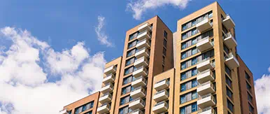 This image features a modern residential apartment block with clean architectural lines, spacious balconies, and large windows set against a bright blue sky. The upward perspective highlights the scale, quality, and contemporary design of urban living spaces.