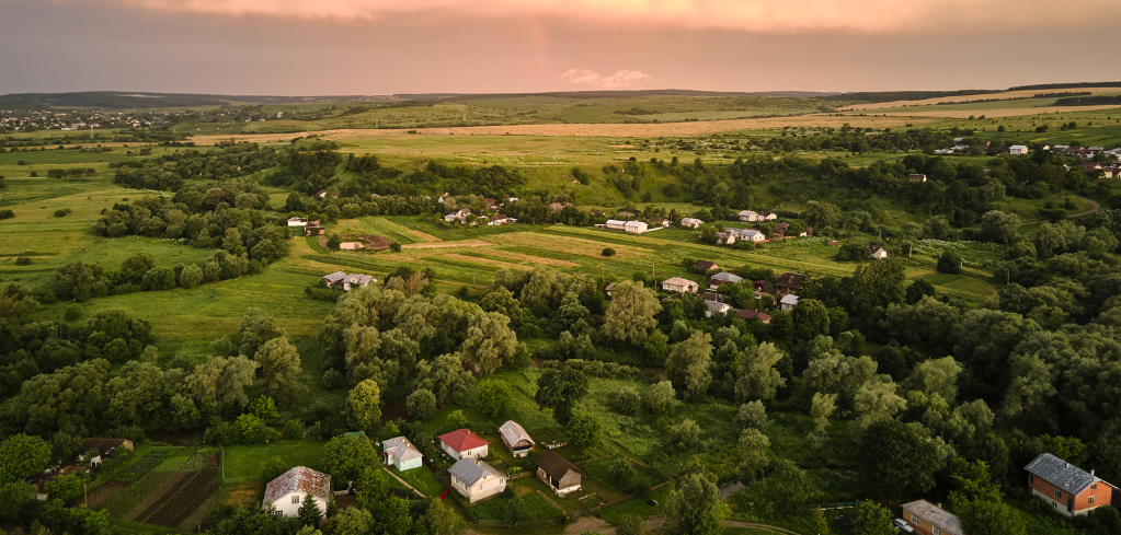 Aerial view of a rural village with scattered houses among green fields and trees under a sunset-tinted sky.