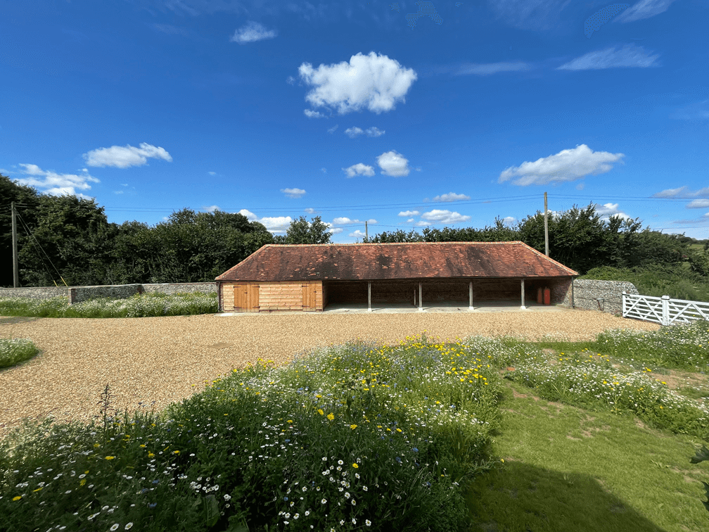 Rustic wooden barn with red-tiled roof surrounded by wildflower meadows and gravel courtyard under bright blue sky.