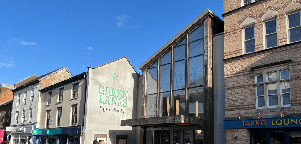 Exterior of Green Lanes Shopping Centre in Barnstaple on a sunny day, with blue skies and neighboring brick buildings.