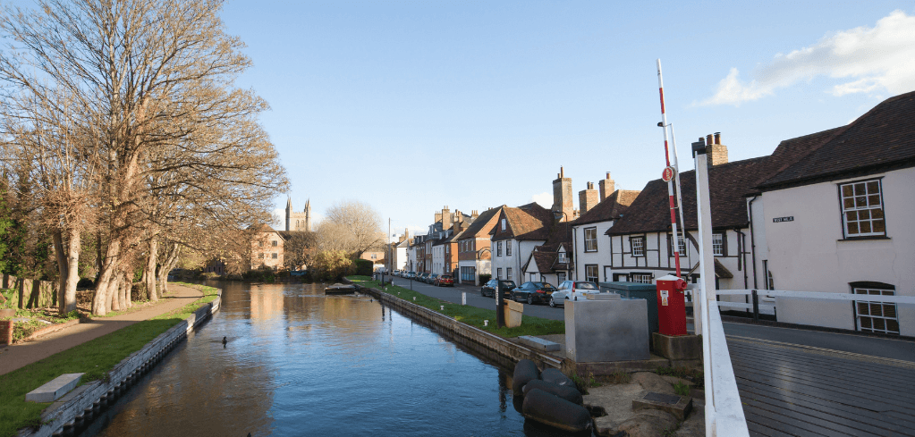 Canal flowing through a quaint English village with historic buildings, bare trees, and a red mailbox under blue sky.
