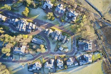 An aerial view of a residential neighbourhood showing multiple houses, winding roads, and surrounding open land on a clear day.