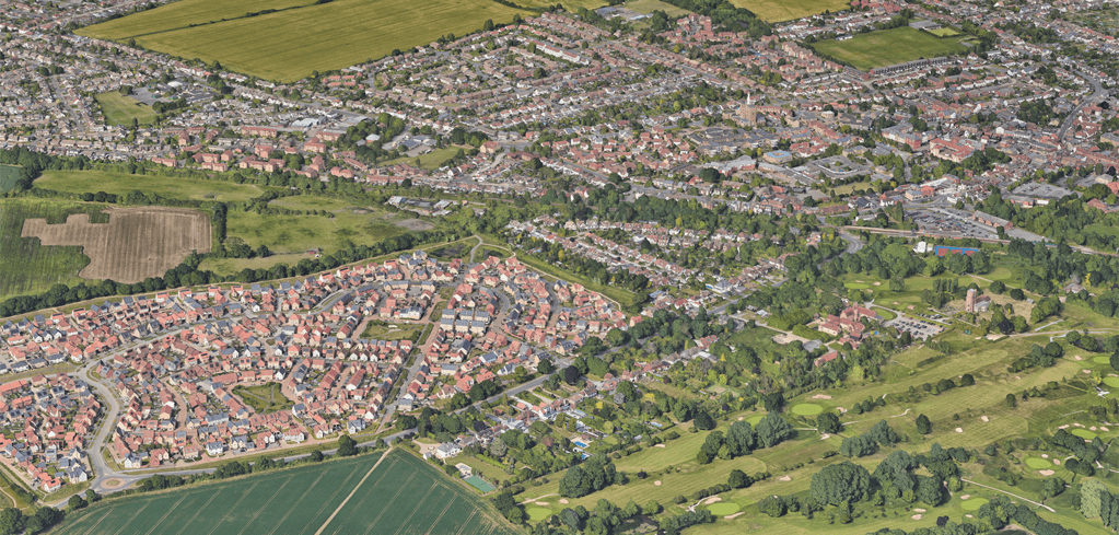Aerial view of a suburban town with red-roofed houses, green fields, tree-lined streets, and a golf course in the foreground.
