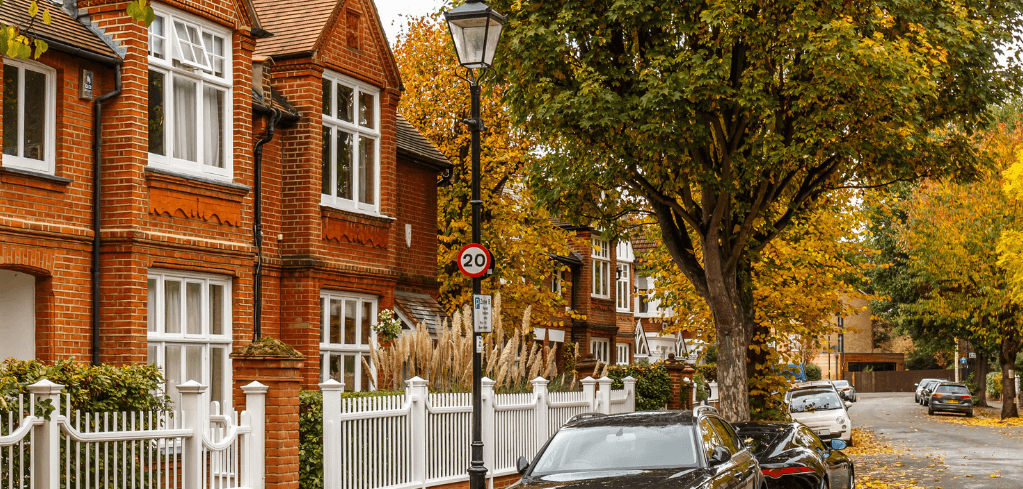 A quiet London residential street in autumn, featuring red brick houses, a 20mph sign, parked cars, and golden-leaved trees.