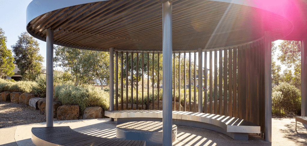 Modern circular pavilion with wooden slat walls and metal columns in a landscaped garden area with sunlight filtering through.